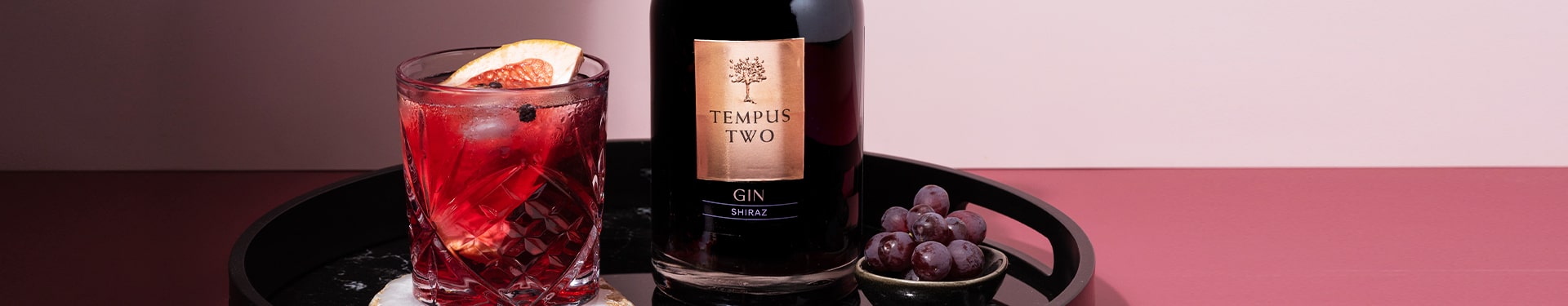 Bottle and glass of Tempus Two Shiraz gin garnished with an orange, next to a bunch of grapes